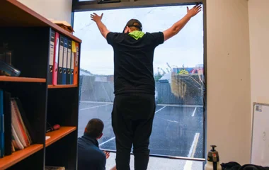 A technician at South West Windscreens & Tint stands at the top of a large office window with arms outstretched, carefully positioning a large sheet of professional window tint in a Busselton office.