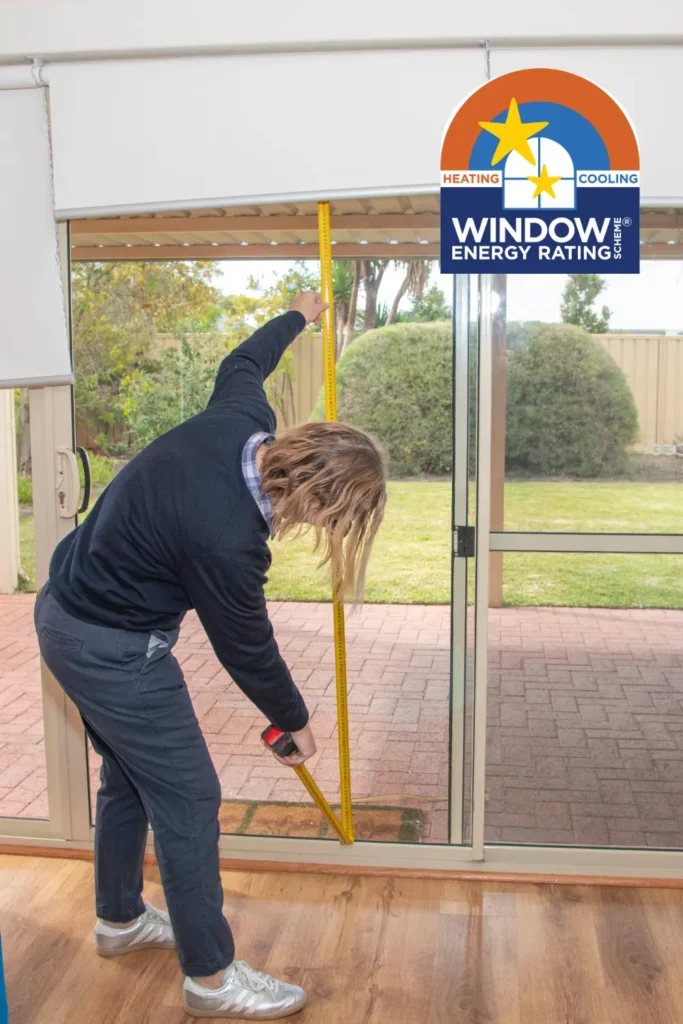 South West Windscreens & Tint team member measuring the height of a large sliding glass door inside a Busselton home using a tape measure, with the official Window Energy Rating Scheme logo overlaid in the upper right corner.