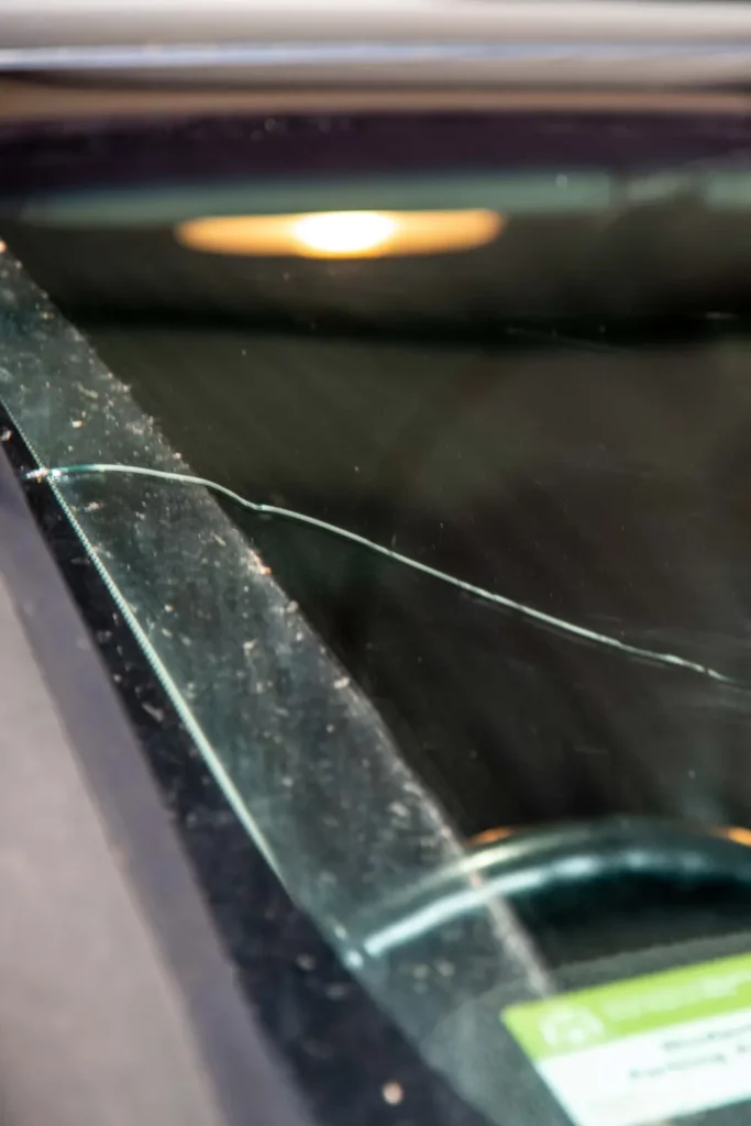 Close-up of a crack running from the lower edge of a vehicle windscreen frame toward the glass surface, showing edge damage that typically requires full windscreen replacement rather than repair.